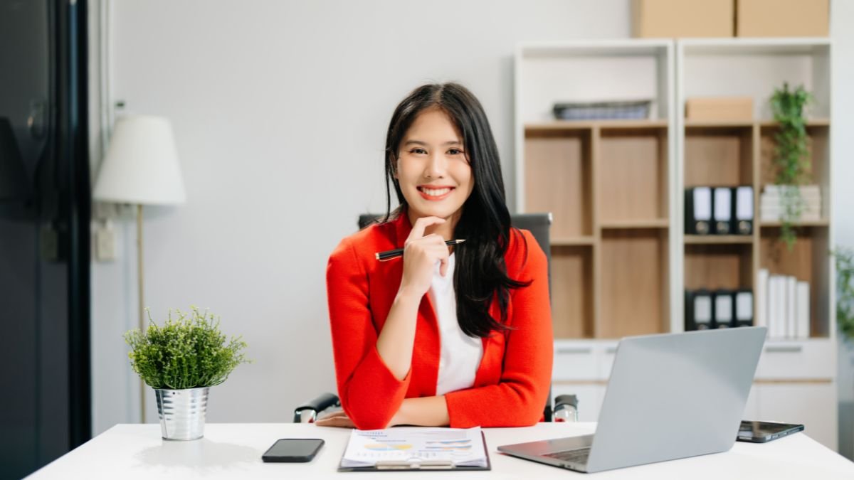 Egy fiatal nő ül egy modern irodában, mosolyogva, kezében tollal.