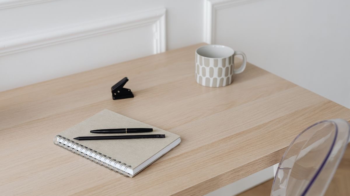 Egy világos fa íróasztalon egy jegyzetfüzet, toll, tűzőgép és egy bögre található.